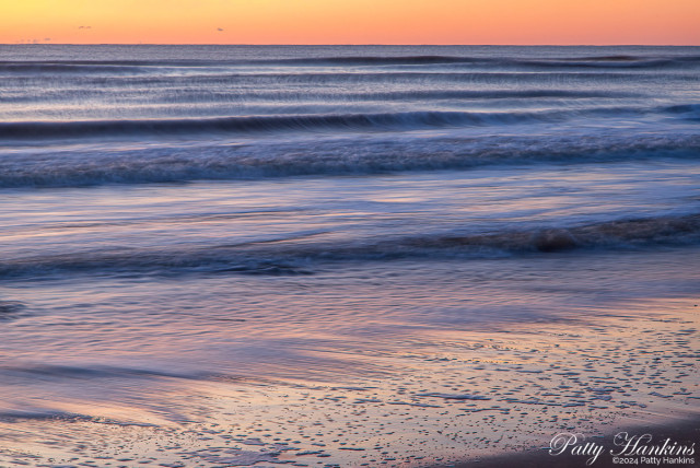 Sunrise at the beach - top of photo is orange and pinks of sunrise, middle is shades of blue in the water with reflections of pinks and oranges, and just hint of sand in the lower right corner