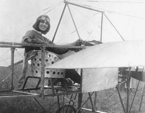 Harriet Quimby sitting in her Blériot XI monoplane. It is an open frame with a metal chair bolted to it. She is dressed for flight with goggles and gloves. She is a white woman with dark hair.
