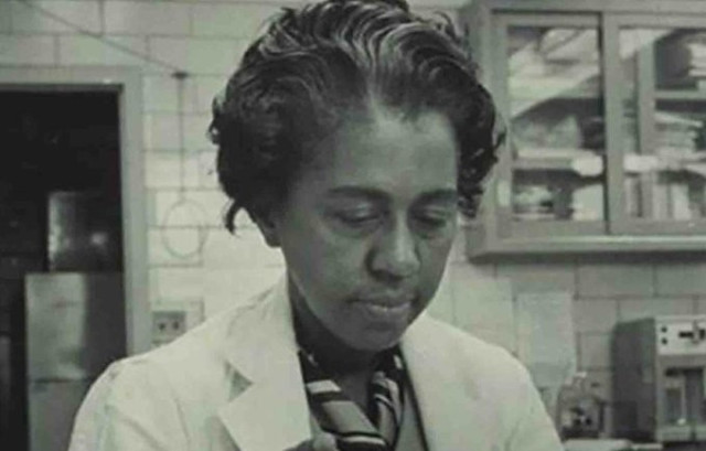 Black and white photo of Marie Maynard Daly in a laboratory with white tile walls and glass-fronted shelves on the wall. Her wavy hair is swept back and she wears a striped kerchief around her neck, under her white lab coat. She is looking down at something on a bench or table in front of her. Lab equipment, including possibly an autoclave, is visible against the wall behind her.