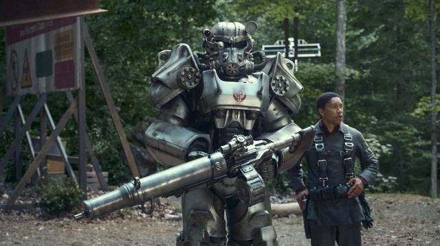 Ein Mann in taktischer Ausrüstung steht neben einem großen, gepanzerten Roboter mit einer Minigun in einem Waldgebiet.