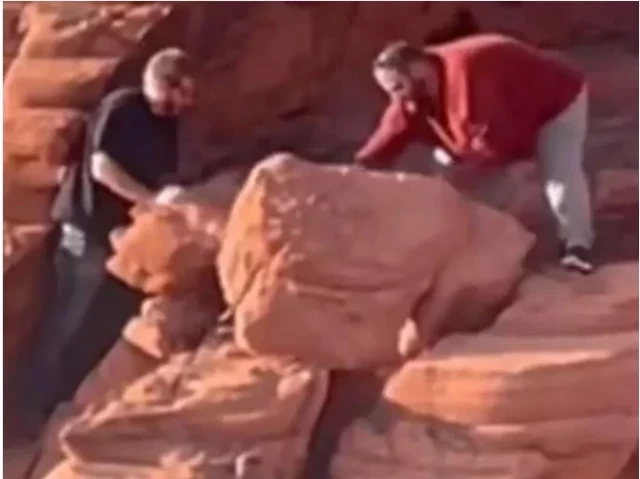 Two men on a red sandstone cliff are toppling a large boulder over the edge. The man on the right looks to be very heavy set and is wearing a long sleeve red shirt, light blue pants, and tennis shoes. He has short brown hair and a beard. He is bent over trying to push a large boulder over the cliff's edge. The man on the left is dressed in black with a short-sleeved tee-shirt. He appears to be loosing a second boulder.