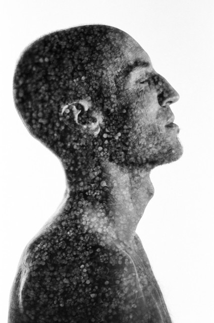 A naked man is standing with his profile to the camera, eyes closed. His skin is mottled by tiny leaves from the double exposure, making it look like a pointillist painting. Black and white. 