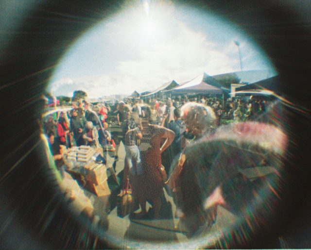 Two (accidentally?) superimposed photos of a mildly chaotic summery Farmers' Market in Dunedin. In one of the exposures, Saira is centred, but with all the crowd shapes you can't really make her face out. There is solar flaring.