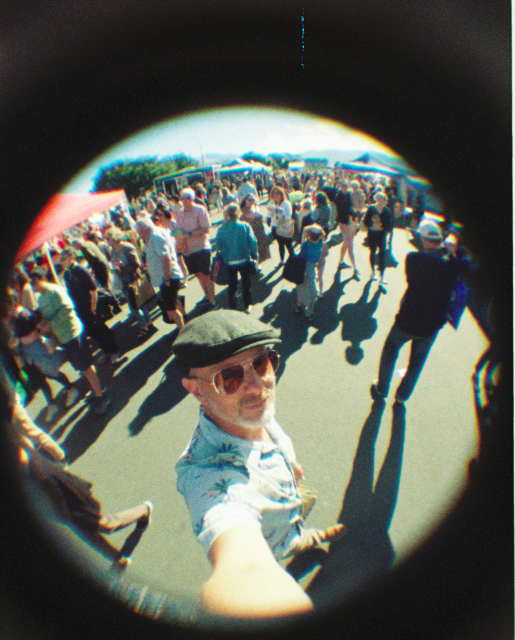 A selfie of me (do I need to say that?) at the market, wearing a green hat and sunglasses, surrounded by people who are small due to the wide FoV of this camera