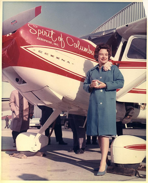 Jerrie Mock in a publicity photo before the flight. She is standing in front of a red and white prop plane called the "Spirit of Columbus". She is wearing a nice coat and matching high heel shoes. She is a white woman with dark hair.
