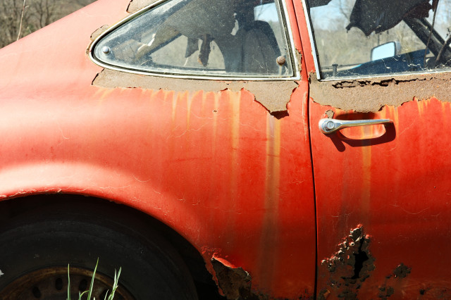 close up side view of a rusted red sports car – peeling – streaking – bubbling metallic paint – hard sunlight – view through the car windows to the driver side rearview mirror – sprigs of grass growing up before the darkness of the wheelwell
