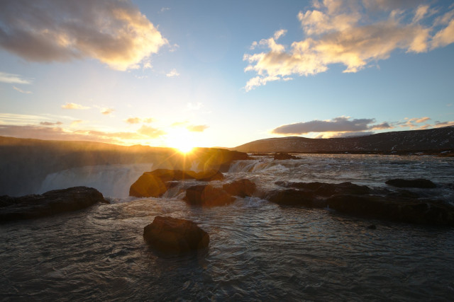 Godafoss waterfall, captured with a wide angle lens. You can see multiple rocks in the water, and the Orange sun creates nice orangey glares in the lens.