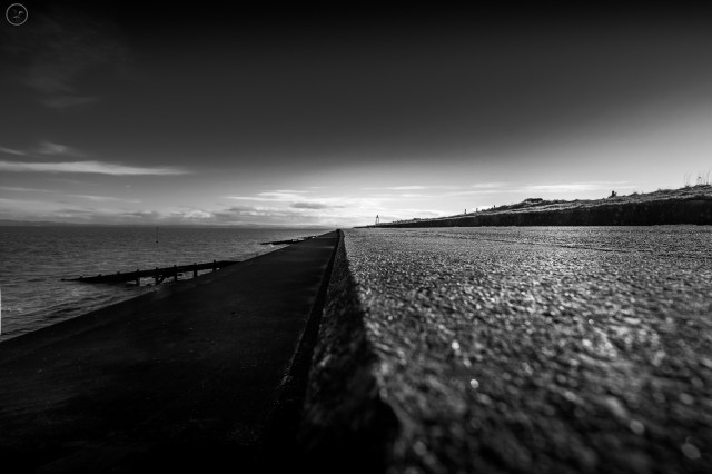 Monochrome shot of concrete shoreline defences with cloud streaked sky above. Sea to background left