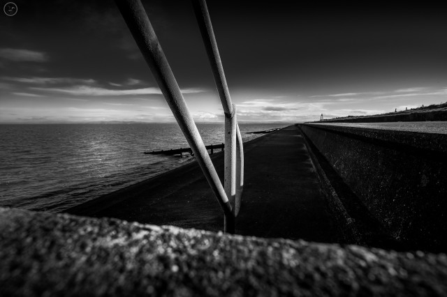 Monochrome shot of concrete shoreline defences with metal railing in foreground and cloud streaked sky above. Sea to background left