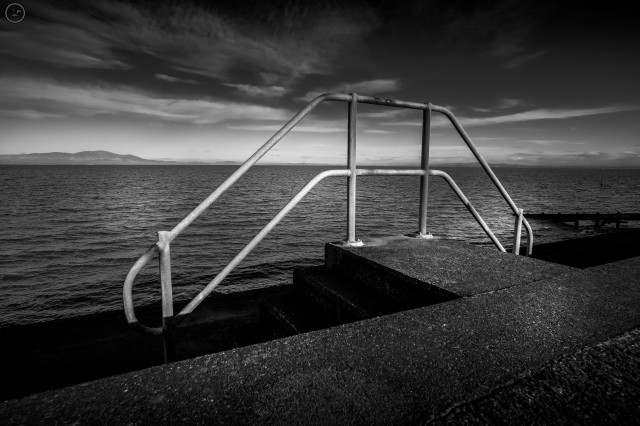 Monochrome shot of concrete shoreline defences with metal railing in foreground and cloud streaked sky above. Sea to background 