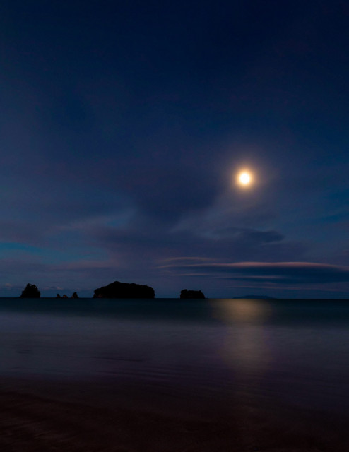 This image is a colour photograph taken at night on a darkened beach with tranquil ocean, with the black silhouette of islands offshore, as a yellow moon rises to reflect in the sea and hang above banks of pink and purple tinged cloud in a vast and clear night sky. 