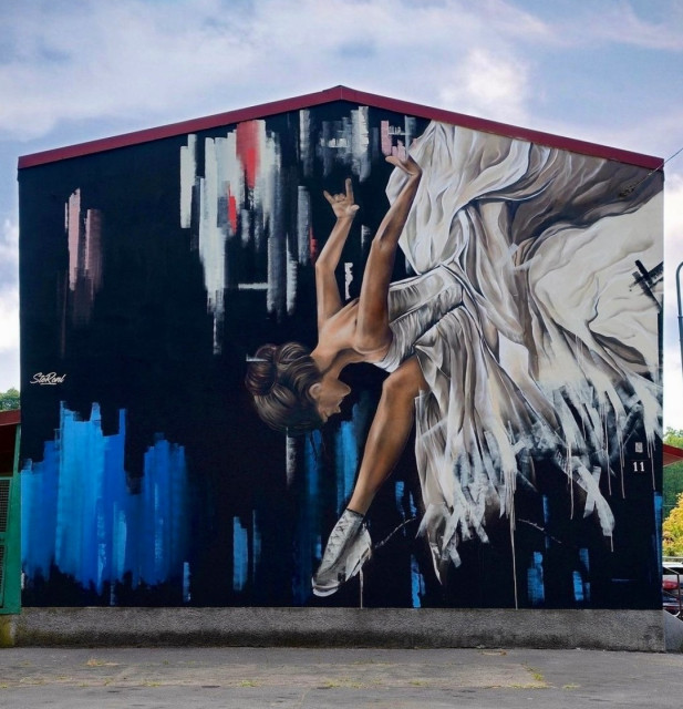Streetartwall. On the outer wall of a one-story building is sprayed / painted mural of a figure ice skater, in the style of an oil painting. The background is painted with dark blue and solid stripes in light blue and white, indicated as an ice landscape. The main figure is a blonde figure ice skater in a white dress, skating powerfully with one leg on the ice, keeping her head lowered and her arms stretched far back. The dress blows in waves behind her, emphasizing the movement.
Info: The muralist has designed together with others for the 2026 Olympics in Milan.