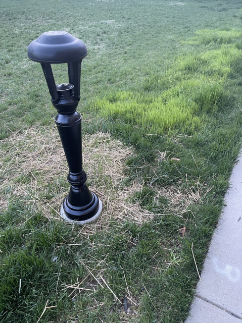 Picture of a light bollard in the grass next to a sidewalk. It's about hip to chest level.