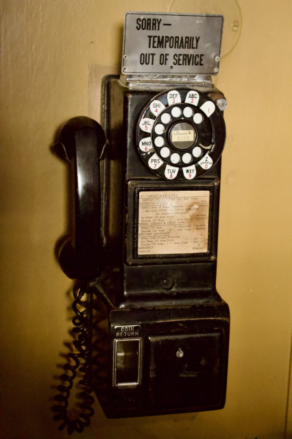 On an amber-colored wall, a very old payphone is mounted. It is dark brown, well worn, with a rotary dialer and a yellowed informational paper placard in the front of it. It has a small steel panel at the bottom with COIN RETURN printed in the steel frame above it.

There is a silver metal sign attached to the top of the phone which reads in black block letters:

SORRY-

  TEMPORARILY 

OUT OF SERVICE


