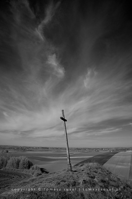 Black and white photograph of a leaning old wooden cross. It stands alone on a hill above the fields, as if watching over them. The clouds in the sky form thin, twisted streaks, contrasting with the dark sky.
