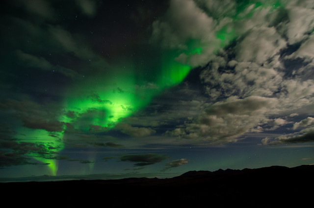 A vivid display of the aurora borealis arcs across the night sky above the dark, silhouetted landscape of the White Mountains in the Alaska Interior. Swirls of bright green Northern Lights contrast with the scattered clouds that are faintly illuminated by the moon's light. Stars dot the dark patches of the sky. The horizon line is defined by the outlined shapes of the mountains, enhancing the natural drama of the auroral light show overhead.