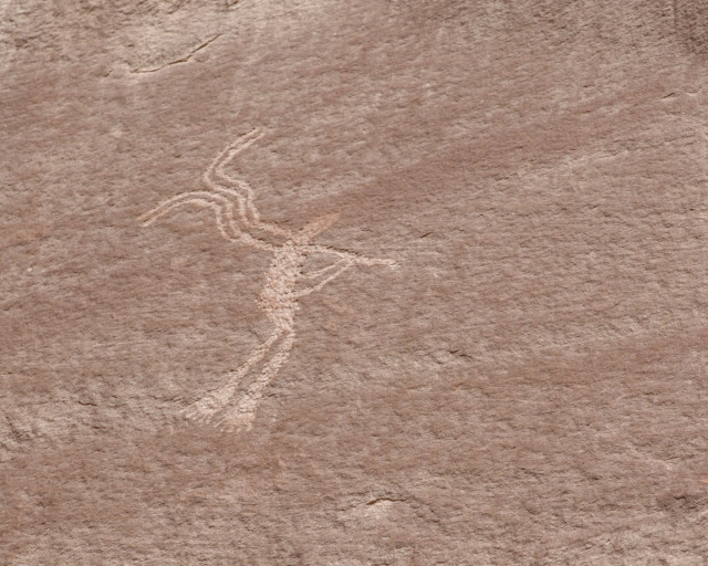 A color photo of what appears to be a human figure holding a long straight object to its mouth with both hands. The two feet are very large showing five toes each. There are two dual curving lines emanating from the figure's back.
