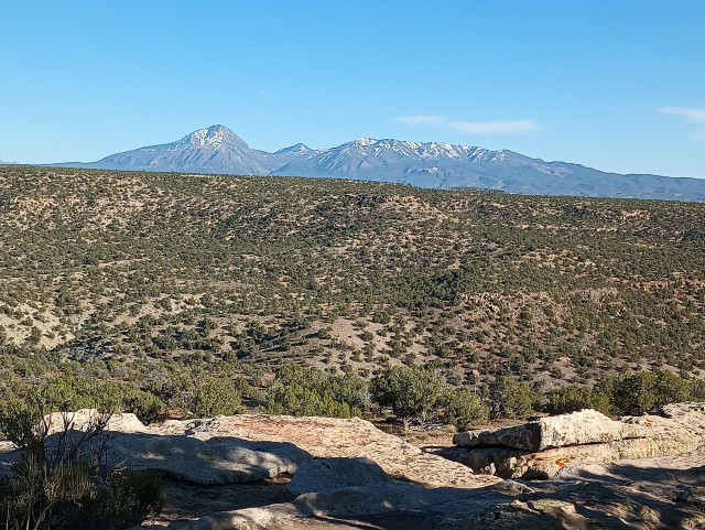 Color photo of a distant mountain with a foreground rolling hill cover in short juniper trees. The profile of the mountain looks like someone laying on their back. The head / nose is on the left. The mountain has small amounts of snow on it.