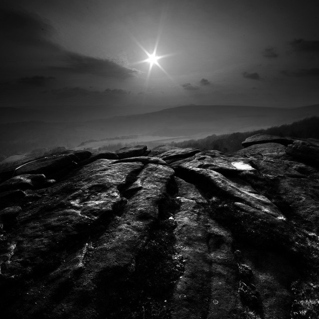 Black and white photo of sunlit rocks in the foreground, with a starburst sun setting over hills beyond.