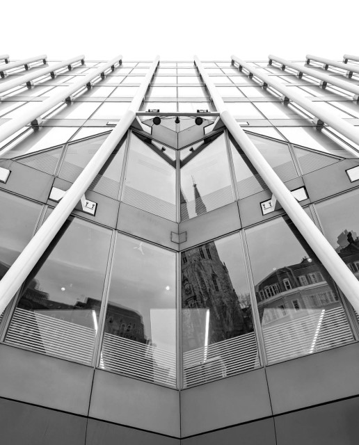 Wide angle black and white photo of a part of the front of a contemporary office building. The face of the building is all windows with vertical metal poles attached at intervals. In the bottom half of the picture the windows angle away from the viewer, with the metal element making a kind of upside-down 'Y'. The older buildings on the other side of the street, including a church, are reflected in these windows, while the ones above reflect just the bright sky.