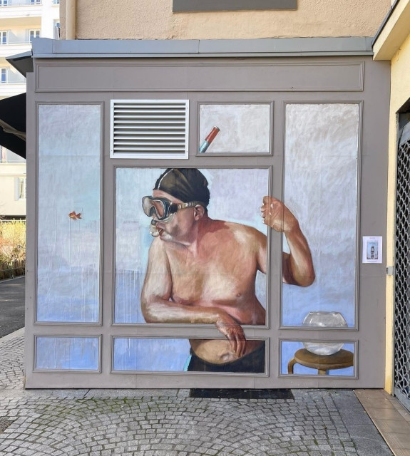 Streetartwall. A whimsical paste-up of a man with diving goggles has been painted on the small exterior wall of a store. The picture shows an older gentleman in black swimming trunks, with diving goggles and a snorkel, looking out of a (painted) window. A tiny red fish swims across the room on the left. Next to him on a small wooden table is a goldfish bowl.
Info: The pasteup was made for a solo exhibition of the artist on site
Galerie Catherine Pennec) in April 2024.