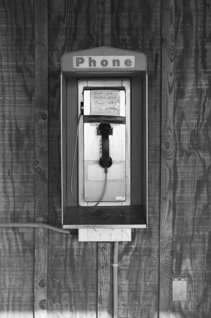 Black and white photo of a pay phone. Sign says "Phone" and there is a black plastic phone hanging on a dial pad made out of metal.