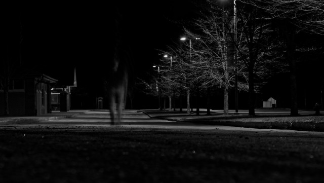 Black and white image of a park at night with street lamps illuminating bare trees and a path, including a building in the background. There's a blurry figure in motion on the left side.