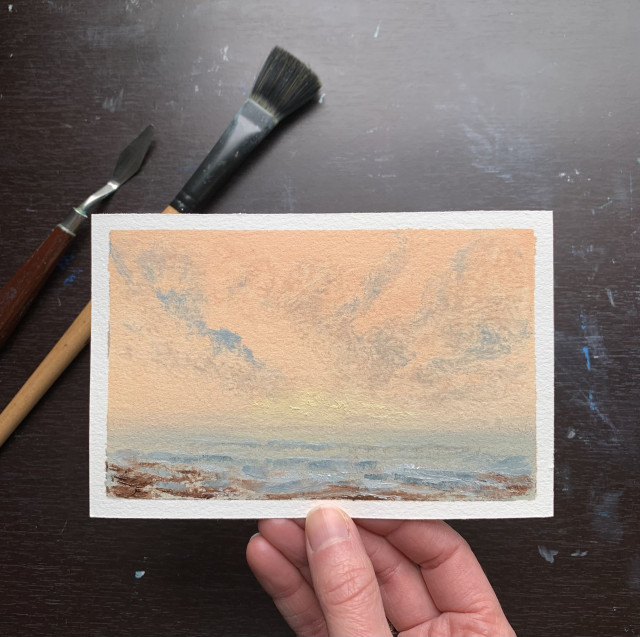Photo of a hand holding an original seascape oil painting by Tisha Mark, "Bright Spring Morning" 4"x6" oil on paper (2024). Seascape painting of a sunrise in a peachy orange sky with some cloud formations. A calm sea meets a shoreline. A palette knife and a paintbrush are visible in the background, behind the upper left corner of the painting.