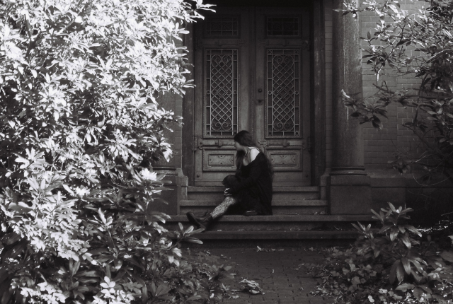 Black and white film photo of a long-haired woman sitting on the steps of a chapel or kind of crypt. Her face is half-turned away from the camera.