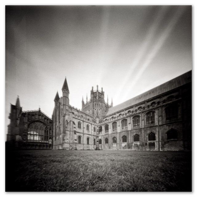 Square black and white pinhole camera photography of Ely Cathedral. Centered on the tower with the nave and transept stretching to either side. Low sun and high clouds give shadows on the underside of the clouds looking like rays radiating from the tower.