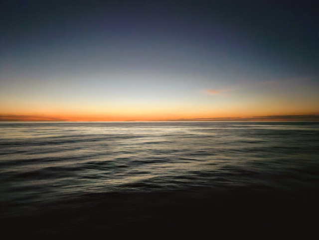 Dark, early morning view across the waves of the Atlantic Ocean beneath a blue sky with the glow of an approaching sunrise casting shades of yellow and orange across the horizon.