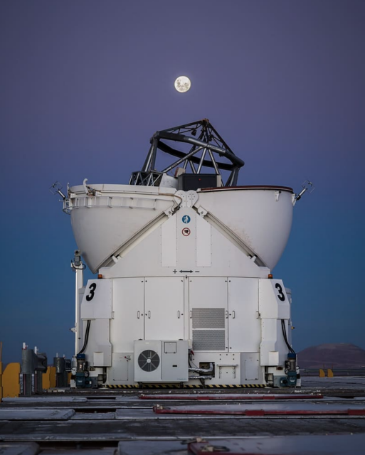 The frame of a telescope sticks out of a white structure shaped like an eggcup, pointing up to the sky. The full Moon is in the background against a dark blue/purple sky.