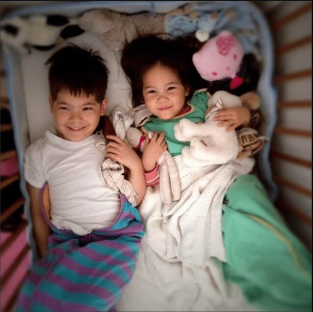 Two siblings, one 5 y/o and one 2 y/o, lying next to each other in a bed, looking up at the camera.