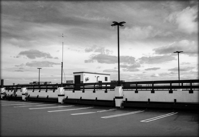The top outside level of a parking garage with tall light poles. Large clouds fill the sky in this black and white picture