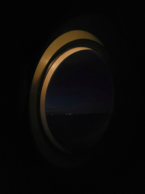 View through a porthole. It is dark inside and you can only see the parts of the porthole that are illuminated by an outside lamp. Outside, you can barely see the mainland against the last light of day on the horizon