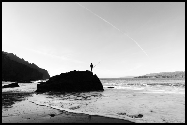 A monochrome photo of a fisherman  - silhouetted, with pole extended - standing on a 20-foot-tall rock on the edge of the water. Waves wash across the beach, surrounding the base of the rock. In the distance, the cliffs of Marin County, overlooking the Golden Gate Channel.

Taken at China Beach.