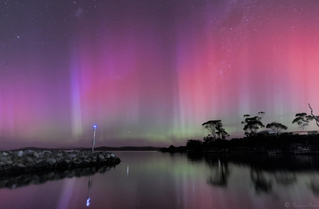 An aurora fills the night sky in the upper 2 thirds, smooth water reflecting the colours in the foreground.  An embankment with a few trees in silhouette to the right, a stone groyne and warning light at left.  The aurora is banded horizontally, green close to the horizon, a pastel red and lighter pink above; and also vertically with columns appearing in the colour. One column has a more blue violet shade in this image.