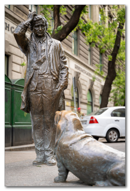 Bronze statue of a man tipping his hat with a dog in the foreground, on a city street.