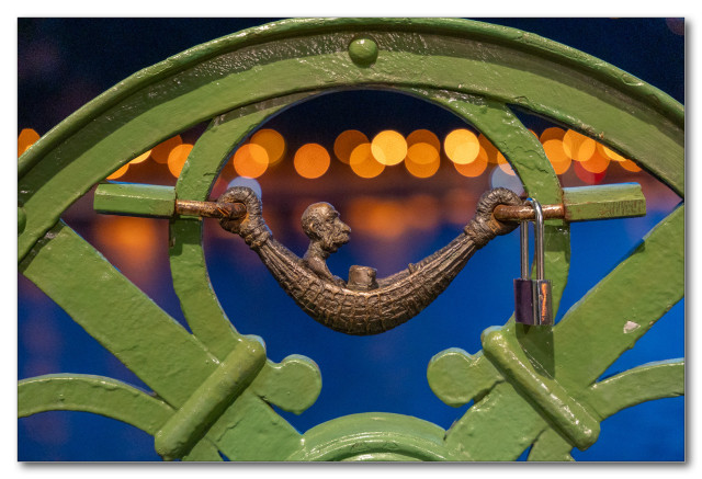 A decorative metal figure in a hammock within a circular frame, part of a green ornate railing, with blurred lights in the background.