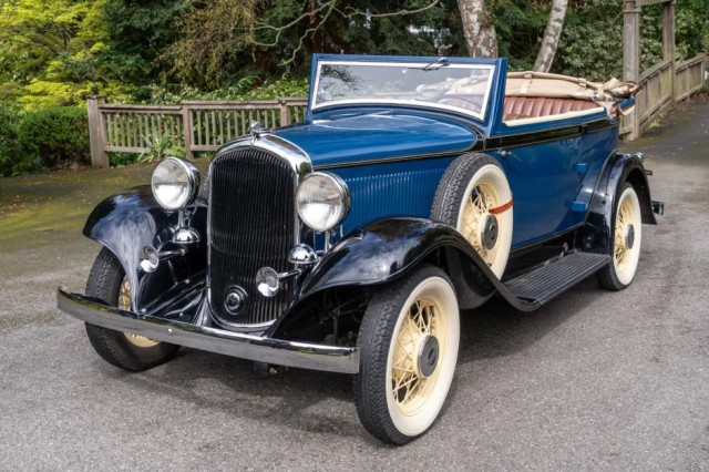 1932 Plymouth Model PB Two-Door Convertible Sedan. An old pre-war-style car with a long nose and tall grille, two rows of bench seating, and an open top. This one’s blue.