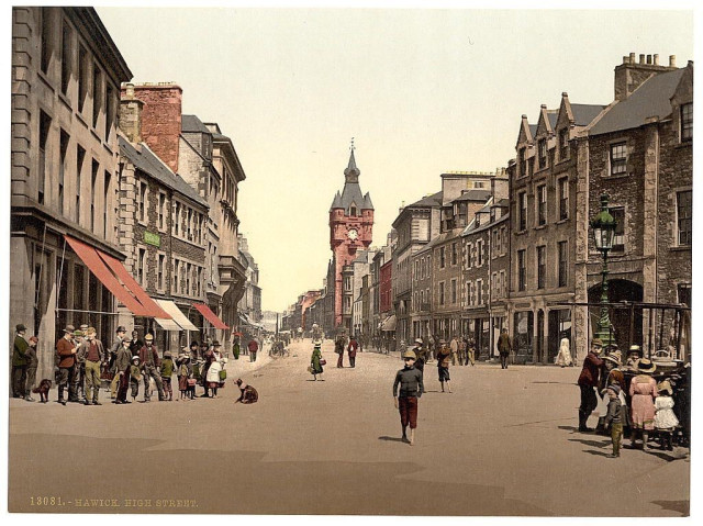  The image is a historical photograph of Hawick's High Street, captured between 1890 and 1900. The street is bustling with activity: people are seen walking along the sidewalk, while others are on horseback and carriages, suggesting a time when these were common modes of transportation. The architecture reveals a mix of styles and materials typical of the era, including stone buildings, some adorned with intricate details and ornate designs. A church steeple is visible in the background, indicating a central role for religion in the community. Overall, the image provides a glimpse into the daily life and urban landscape of Hawick during the late 19th century.