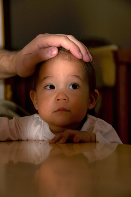 A 1-year old baby holding a table. Her hands and face is visible above the table. Her fathers hand is on top of her head, caught in mid action, but also shows the scale.
