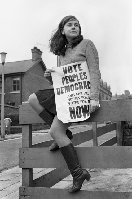 Bernadette Devlin campaigning for election. She's a white woman sat on a fence and holding a poster reading "vote people's democracy - jobs for all - houses for all - votes for all"