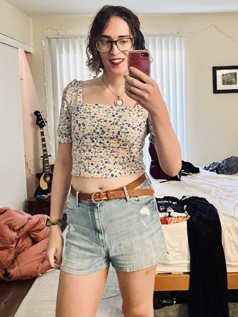 Myself, a white trans woman with wet brown hair taking a selfie while wearing a square-necked floral croptop, denim shorts, and a brown belt