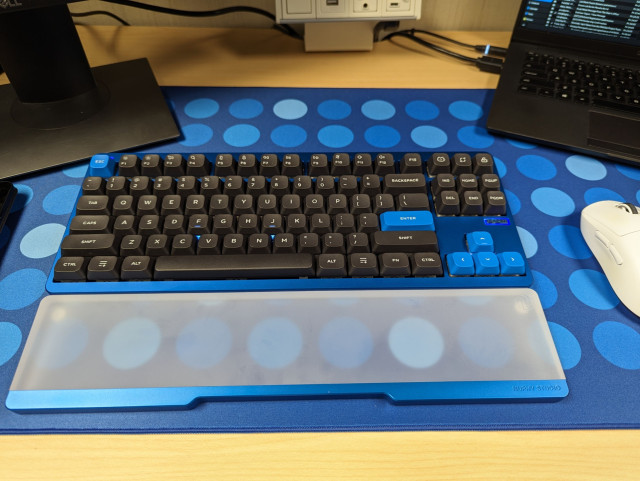 a keyboard with blue base and black keycaps. in front of the keyboard is a glossy transparent wrist rest. they are on a desk with a desk mat that is the same color as the keyboard 