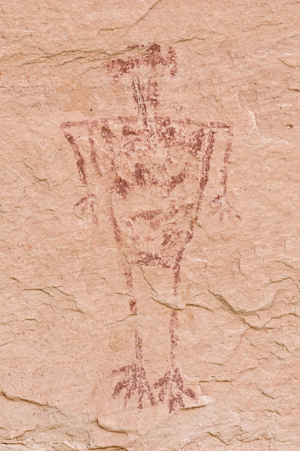 A prehistoric dark red pictograph on lighter red sandstone. It's an anthropomorph. The body is triangular shaped. The arms hang down from wide shoulders with hands spread wide. At the end of long stick like legs are bird like looking feet. The head is hard to see but it is kind of like a triangle with a mob of hair on top.