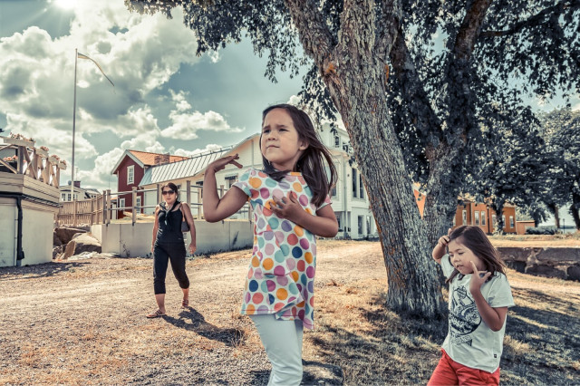A photo of a woman and 2 children walking in a seaside town. There is a tree in the centre, but the people in the image appear unware of that a photo is being taken.