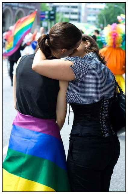 Ein Paar küsst sich. Eine der Personen hat eine Regenbogenflagge um die Hüfte geschlungen. Die andere Person trägt eine karierte Bluse und ein schwarzes Korsett. Im Hintergrund weitere Menschen mit Regenbogenfarben.
