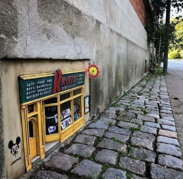 A teeny, real-looking record store front built onto a grey wall along a cobblestone street. It's even been given a Banksy touch with some spray paint!