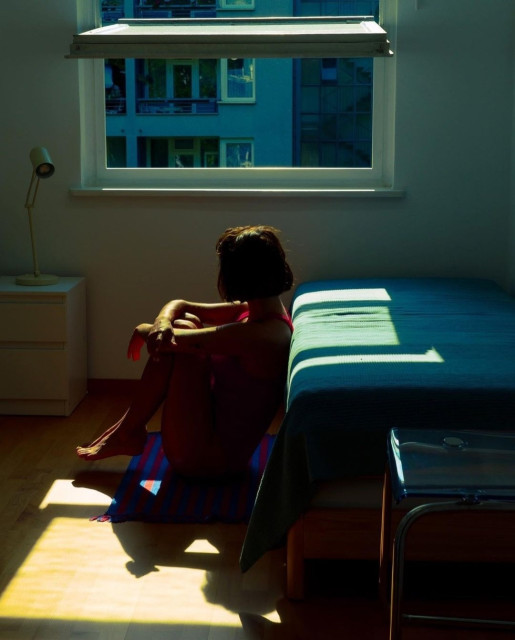 Photography. A color photo of a young woman sitting in front of an open window. The room is bathed in a twilight of blue/green and a little red. A young woman with short black hair and a red summer dress sits on the light-colored floor in front of her bed. She has her arms on her knees and is looking towards the window and the sunlight streaming in. Shadows and light dominate the photo. The room has a light blue bed and a simple metal stool on the right and a white bedside table on the left next to an open tilt window in the middle. The view is of a house front opposite. The photo has a calm atmosphere and is cool and warm at the same time. Between solitude or enjoying the sunlight in the shade, it is up to the viewer to interpret the photo for themselves.